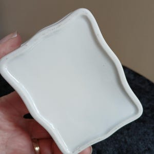 May include: A white ceramic dish with a unique, wavy edge. The dish is held in a hand, showcasing its smooth, glossy surface. The design is simple and elegant, suitable for various uses.