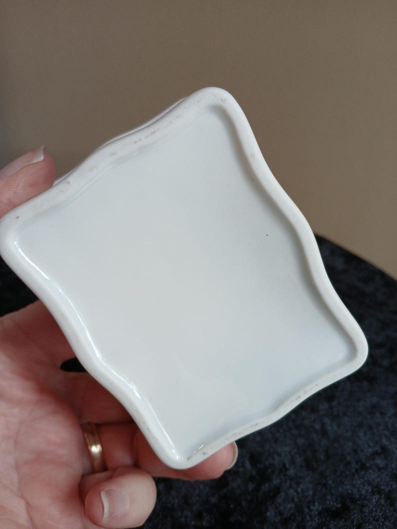 May include: A white ceramic dish with a unique, wavy edge. The dish is held in a hand, showcasing its smooth, glossy surface. The design is simple and elegant, suitable for various uses.