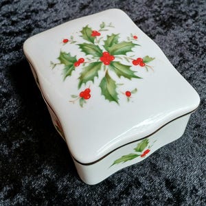 May include: A white ceramic trinket box with a lid, featuring a holly and berry design. The box has a curved shape and a brown trim. The holly leaves are green, and the berries are red.