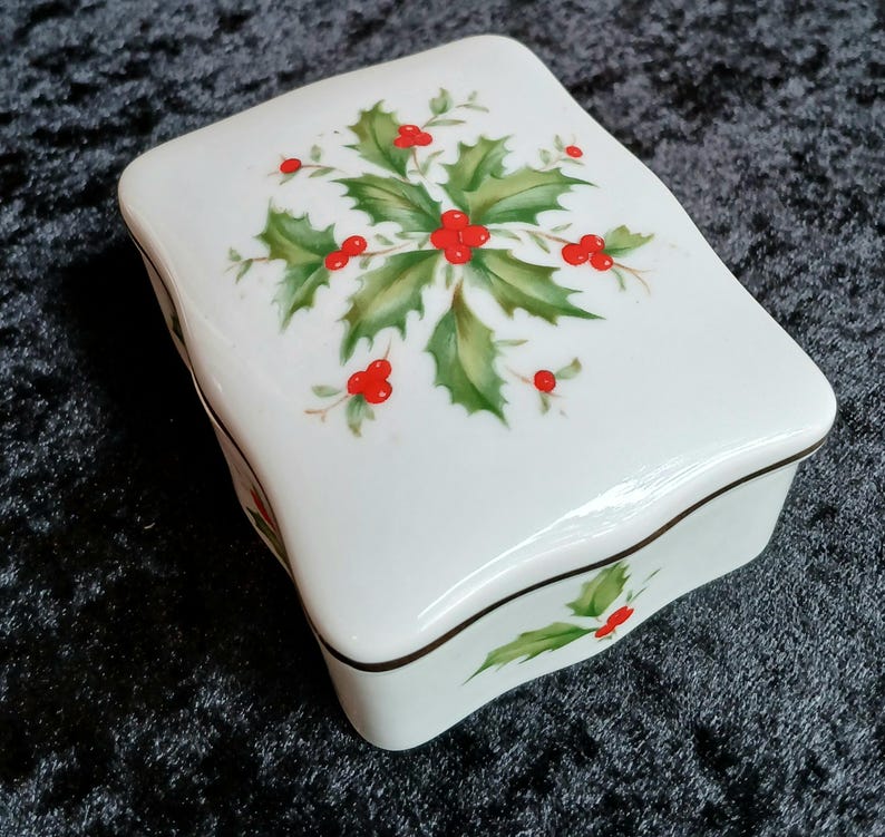 May include: A white ceramic trinket box with a lid, featuring a holly and berry design. The box has a curved shape and a brown trim. The holly leaves are green, and the berries are red.