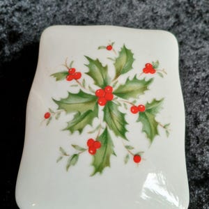 May include: A white, rectangular ceramic dish with rounded edges. The dish is decorated with a central design of green holly leaves and bright red berries, a classic Christmas motif.