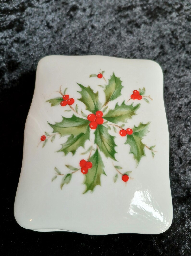 May include: A white, rectangular ceramic dish with rounded edges. The dish is decorated with a central design of green holly leaves and bright red berries, a classic Christmas motif.