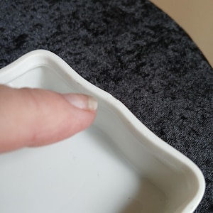 May include: Close-up of a white, square-shaped dish with a wavy edge. The dish is made of a glossy material and has a simple, elegant design. The dish is empty and appears to be clean.