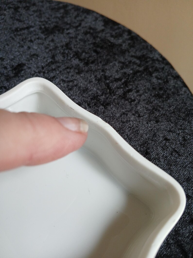 May include: Close-up of a white, square-shaped dish with a wavy edge. The dish is made of a glossy material and has a simple, elegant design. The dish is empty and appears to be clean.