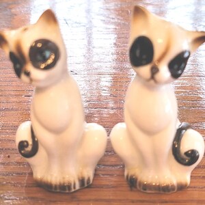 May include: Two ceramic cat figurines. The cats are white with brown ears and black eyes. The figurines have a sitting pose with a curled tail. The base of the figurines is brown.