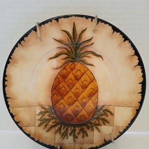 May include: A decorative plate featuring a hand-painted pineapple design. The pineapple is a warm orange color with a textured appearance, set against a beige background with a black rim. The plate has a rustic, farmhouse aesthetic.