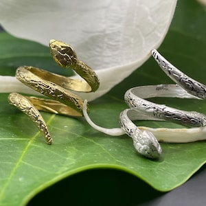 Op de afbeelding: Twee slangenringen, een gouden en een zilveren. De ringen zijn opgerold en hebben een getextureerd oppervlak. De ringen liggen op een groen blad en een witte bloem.