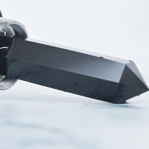 May include: A black, faceted crystal point is partially inserted into a silver-colored metal holder. The crystal has a polished, reflective surface. The background is a light blue and white marbled surface.