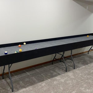 May include: A long, black wooden table with a gray felt surface and a set of billiard balls. The table is supported by two folding metal legs.