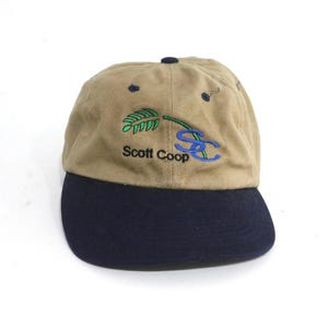 May include: Tan and navy blue baseball cap with an embroidered green leaf and the text "Scott Coop" and "SC" in blue.