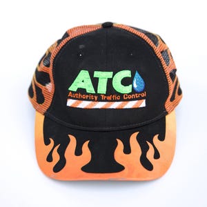 May include: Black baseball cap with orange flame design on the brim. The cap features the text "ATC Authority Traffic Control" in green and white. The top of the cap has an orange mesh design.