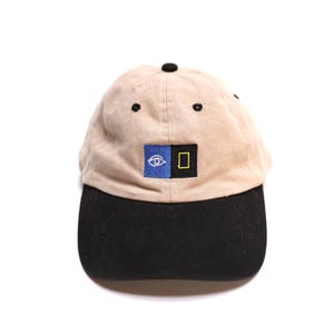 May include: A beige baseball cap with a black brim and a black button on top. The front features a rectangular patch divided into a blue square with a white eye icon and a black square with a yellow rectangular outline, centered on the crown.