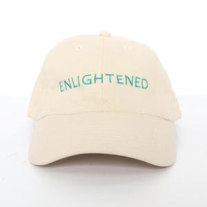 Peut inclure: Casquette de baseball crème avec le mot "ENLIGHTENED" brodé en vert sur le devant. La casquette a une visière incurvée et une couronne arrondie. Le chapeau est fait d'un tissu doux et texturé.