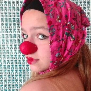 Puede incluir: Una persona joven con una nariz de payaso roja y lápiz labial rosa lleva un pañuelo de cabeza rosa con un patrón floral. El pañuelo está atado en un nudo en la parte posterior de la cabeza. La persona está mirando hacia un lado y tiene el pelo largo y rubio.