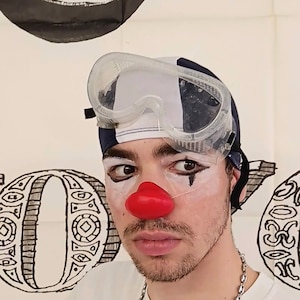 Puede incluir: Una persona lleva una gorra de baño a rayas blancas y azules y gafas de seguridad transparentes. Tiene pintura facial blanca con delineador de ojos negro y una nariz de payaso roja. Lleva una camiseta blanca con un collar de cadena plateada. El fondo es blanco con diseños abstractos en blanco y negro.