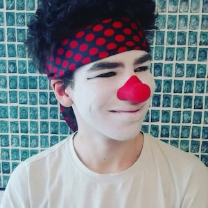Puede incluir: Una persona con maquillaje de payaso con una nariz roja y una diadema de lunares negros y rojos. Lleva una camiseta blanca con una imagen de dibujos animados.