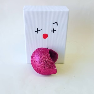 Puede incluir: Una caja blanca con un punto rojo y signos más negros dibujados en ella. Un objeto en forma de corazón rosa brillante se encuentra delante de la caja.