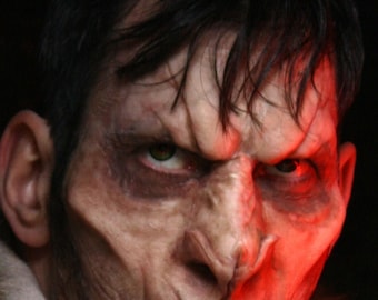 May include: Close-up of a person's face with dramatic makeup, possibly for a horror or fantasy theme. The skin has a pale, almost undead appearance, with dark circles around the eyes and a menacing expression. Red lighting adds to the eerie effect.