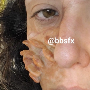 May include: Close-up of a person's face with a special effects makeup application. The makeup is designed to look like a fleshy, textured growth on the side of the face. The text '@bbsfx' is visible on the image.