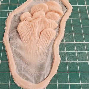 May include: A close-up of a light pink silicone mold of a cluster of mushrooms. The mold is on a green cutting mat.
