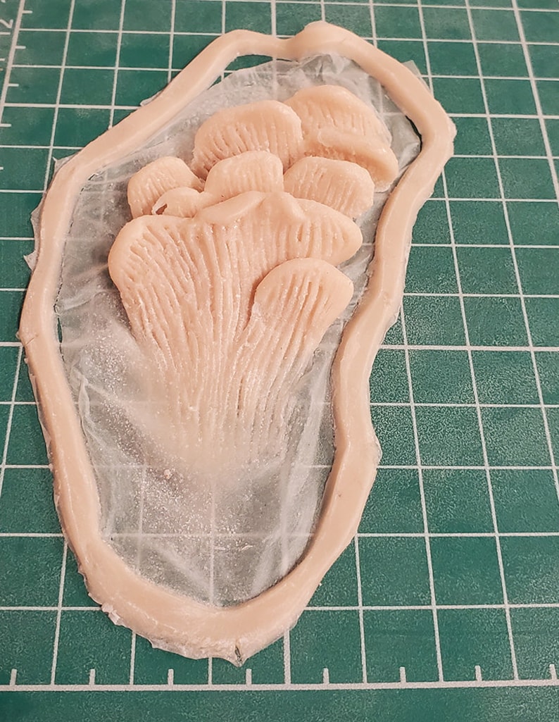 May include: A close-up of a light pink silicone mold of a cluster of mushrooms. The mold is on a green cutting mat.