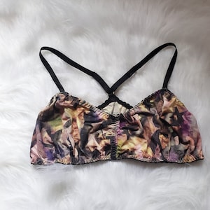 Puede incluir: Un bralette colorido con un estampado floral en tonos morados, amarillos y marrones. El bralette tiene tirantes y ribetes negros. Las correas se cruzan en la espalda. La prenda está hecha de una tela suave y ligera.