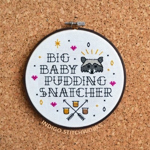 Puede incluir: Aro de bordado blanco con texto negro que dice "Big Baby Pudding Snatcher" con una cara de mapache en el medio. El texto está rodeado de pequeños corazones rosas, estrellas amarillas y diamantes negros. El aro está sobre un fondo de corcho marrón.