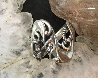 Könnte beinhalten: Ein silberfarbener Ring mit einem verzierten, wirbelnden Design. Der Ring weist komplizierte Muster und Kurven mit poliertem Finish auf. Der Ring wird auf einer weiß-grauen Oberfläche mit natürlicher, steinartiger Textur präsentiert.