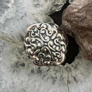 May include: A silver-colored ring with an ornate, swirling design. The ring's surface is covered in detailed, flowing patterns. The ring is set against a backdrop of light and dark gray stone.