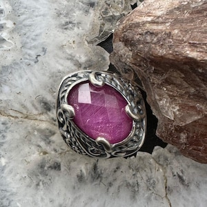 May include: A silver ring with an oval, faceted, magenta gemstone. The ring has an ornate, filigree design around the gemstone. The ring is set against a light gray and brown background.