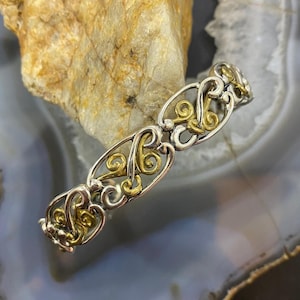 May include: Silver and gold-toned bracelet with an ornate, openwork design. The bracelet features a repeating pattern of swirling shapes and curves, with gold accents. The metalwork is detailed and intricate, creating a delicate and elegant piece of jewelry.