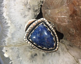 Könnte beinhalten: Ein Silberring mit einem dreieckigen, tiefblauen Lapislazuli-Stein. Der Stein ist in einem dekorativen, strukturierten Silberband gefasst. Der Ring wird auf einer hellgrauen und weißen Steinoberfläche mit einem braunen und klaren Kristall im Hintergrund präsentiert.