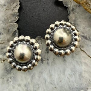 May include: A pair of round silver earrings featuring a smooth, domed center surrounded by a twisted rope border and an outer edge of small, spherical beads. The metal has a polished finish with subtle darkened areas in the recessed details.