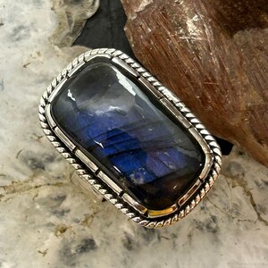 May include: A rectangular ring featuring a polished labradorite gemstone with deep blue and purple iridescence. The stone is set in a silver metal bezel with a decorative twisted rope border design around the perimeter of the setting.