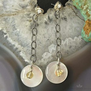 May include: A pair of silver-toned drop earrings featuring a vertical chain link design. Each earring ends in a circular white shell disc with a small, translucent yellow bead suspended in the center. The earrings have a post-back closure.