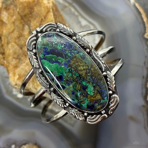 May include: Silver cuff bracelet with an oval, multi-coloured stone in shades of blue, green, and brown. The stone is set in a decorative silver frame with leaf and scroll details. The bracelet has an open design.