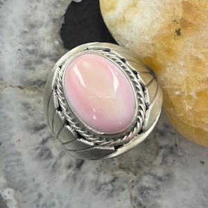 May include: A sterling silver ring featuring a large, oval-shaped, smooth pink stone set in the center. The stone is surrounded by a twisted rope border, and the band is decorated with etched leaf-like patterns on both sides of the setting.