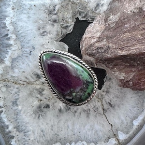 Könnte beinhalten: Ein tropfenförmiger Ring mit einem polierten Rubin-Zoisit-Edelstein mit dunkelvioletten und grünen gesprenkelten Mustern. Der Stein ist in eine silberfarbene Metallfassung mit einem gedrehten seilartigen Randdetail um den äußeren Rand gefasst.