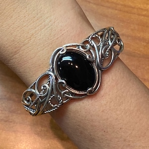 Puede incluir: Brazalete de puño plateado con una piedra negra ovalada y brillante. El brazalete presenta un diseño calado adornado con patrones arremolinados y detalles en forma de cuerda, mostrando una estética vintage. Una joya clásica.