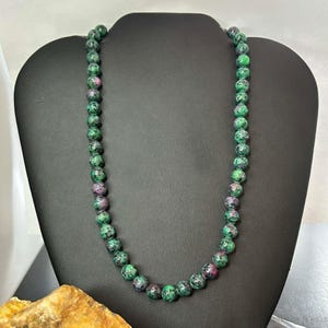 Puede incluir: Un collar compuesto por cuentas redondas y pulidas que presentan un patrón moteado de verde bosque profundo y rosa magenta vibrante. Las cuentas son de tamaño uniforme y están ensartadas para formar un hilo circular continuo de piedra.