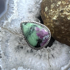 May include: A teardrop-shaped ring featuring a vibrant green and magenta gemstone. The stone is set in a silver-colored metal with a twisted rope design around the edge. The ring is displayed on a white and gray surface.