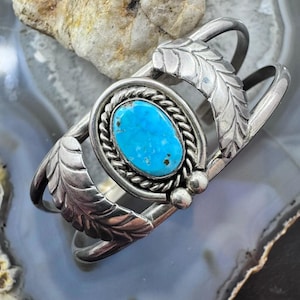 Op de afbeelding: Een zilveren manchetarmband met een heldere, turquoise ovale steen, gezet in een touwachtig kader. De armband heeft bladvormige accenten en een gepolijste afwerking, met een ontwerp uit het zuidwesten.