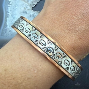 May include: A cuff bracelet featuring a central silver-toned band with a repeating stamped pattern of small bear silhouettes and geometric triangular lines, bordered by two smooth copper-toned edges that frame the detailed metalwork design.