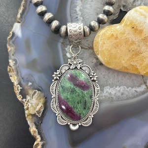 May include: A necklace featuring a large oval pendant with a green and magenta zoisite stone set in a decorative silver frame with floral accents, suspended from a strand of round, polished silver beads with a textured cylindrical bail.