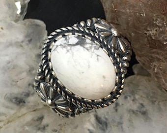 Könnte beinhalten: Ein silberner Ring mit einem ovalen, weißen Stein mit grauer Marmorierung. Der Stein ist in einem dekorativen Silberrahmen mit einem seilartigen Rand und floralen Akzenten gefasst. Der Ring wird auf einer hellen, strukturierten Oberfläche präsentiert.