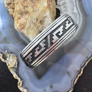 May include: A silver-colored ring with a geometric design. The ring features a series of parallel lines and stylized shapes. The ring is displayed on a blue and gray stone surface, with a light brown rock in the background.