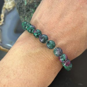 May include: A beaded bracelet with round gemstones in green and red hues. The bracelet is strung on a thin, silver-coloured cord. The gemstones have a speckled appearance.
