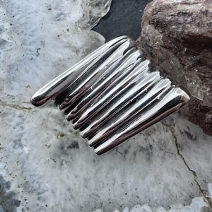 May include: A polished silver ring with a distinctive design. The ring has a series of parallel, elongated, rounded bars, creating a textured surface. The ring is set against a backdrop of light and dark grey stone.