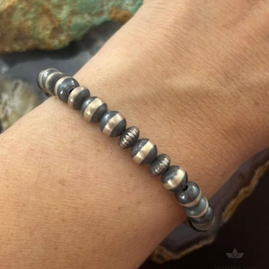 May include: A bracelet with dark grey and silver beads. The bracelet has round beads with a striped design, and smaller, textured beads. The jewellery is displayed on a person's arm. The bracelet is likely made of metal.