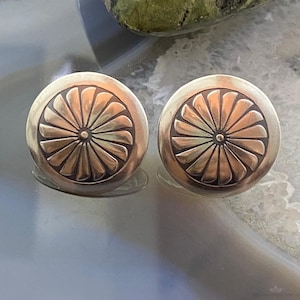 May include: A pair of round, silver-toned stud earrings. Each earring features a raised, sunburst-like design with a central dot. The earrings are displayed on a reflective surface, showcasing their metallic sheen.